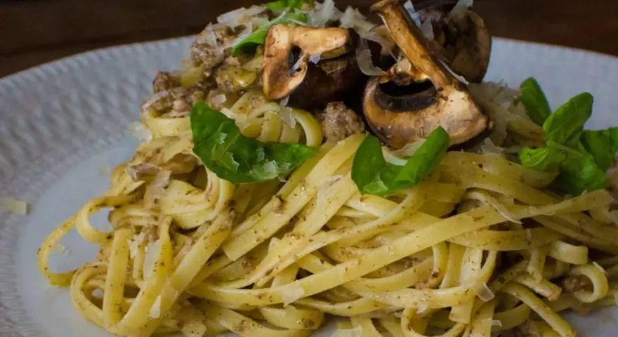 Pasta con champiñones, carne picada y queso rallado, decorada con hojas verdes.