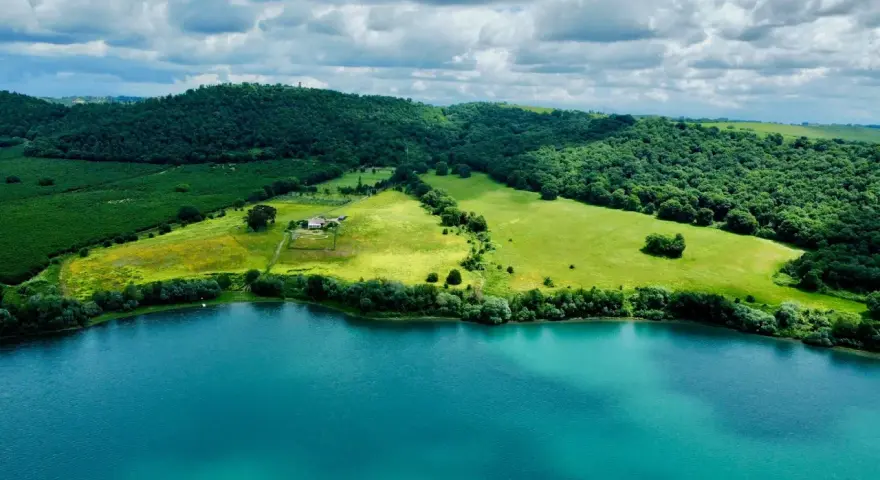 Acque turchesi del lago di Martignano lambiscono dolci colline verdi, con un casale isolato tra i campi.
