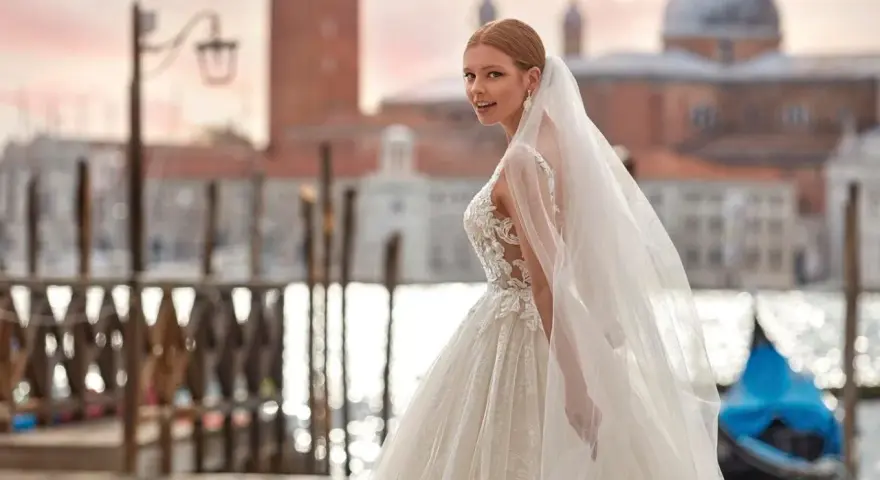 Sposa con abito da sogno e velo svolazzante a Venezia, con gondola blu sullo sfondo. Perfetta per un matrimonio da favola, anche con stivali sposa.