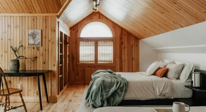 Dormitorio con paredes y techo de madera, cama con mantas y almohadas, ventana arqueada y escritorio.