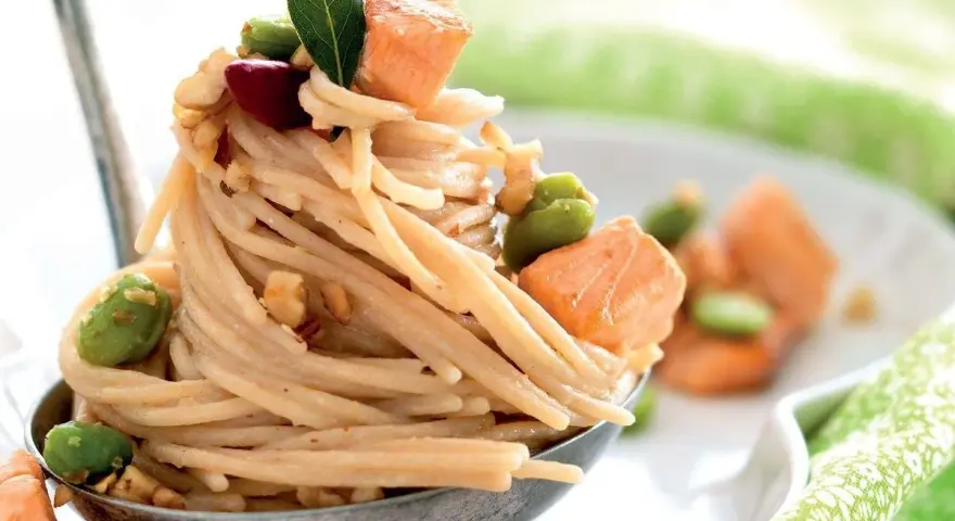 Spaghetti di riso con salmone, fave e noci, guarniti con una foglia verde. Un piatto invitante e colorato.