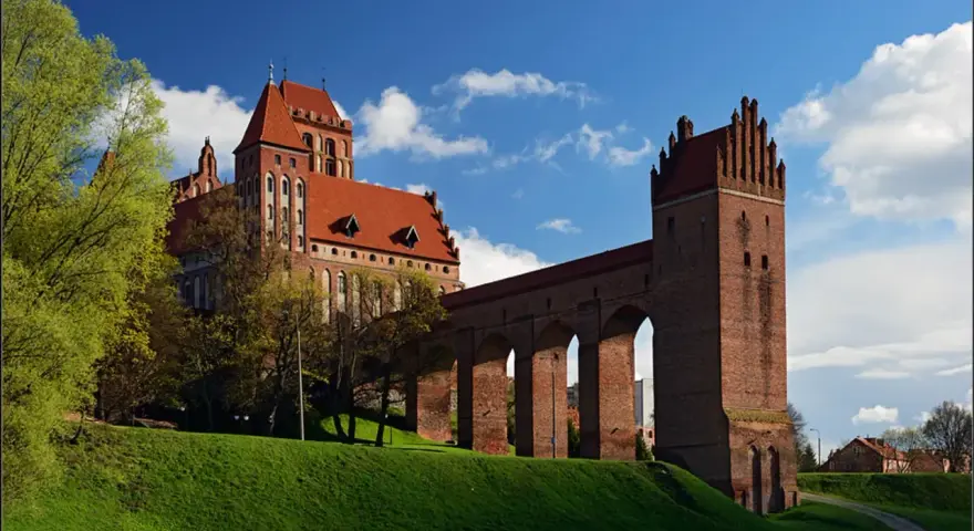 Majestatyczny zamek w Kwidzynie, z czerwonej cegły, z charakterystycznym gdaniskiem i długim, arkadowym murem obronnym, wznosi się na zielonym wzgórzu pod błękitnym niebem.