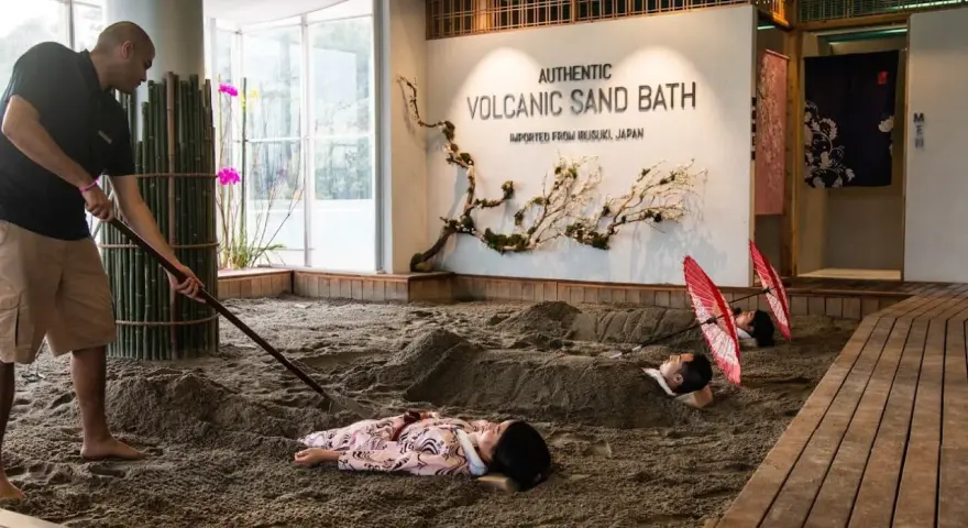 A man rakes sand for a volcanic sand bath. Two people lie buried in the sand, with red umbrellas over their heads.