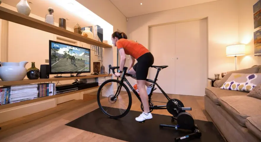 Woman in her home cycling pain cave, training indoors on a bike trainer while watching a virtual race on a large screen.