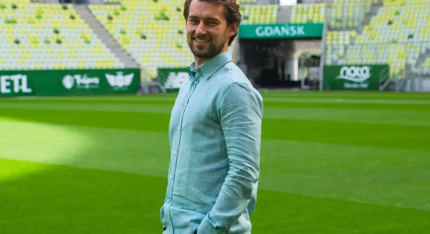 Uśmiechnięty Tomasz Kaczmarek, trener, stoi na tle stadionu w Gdańsku.