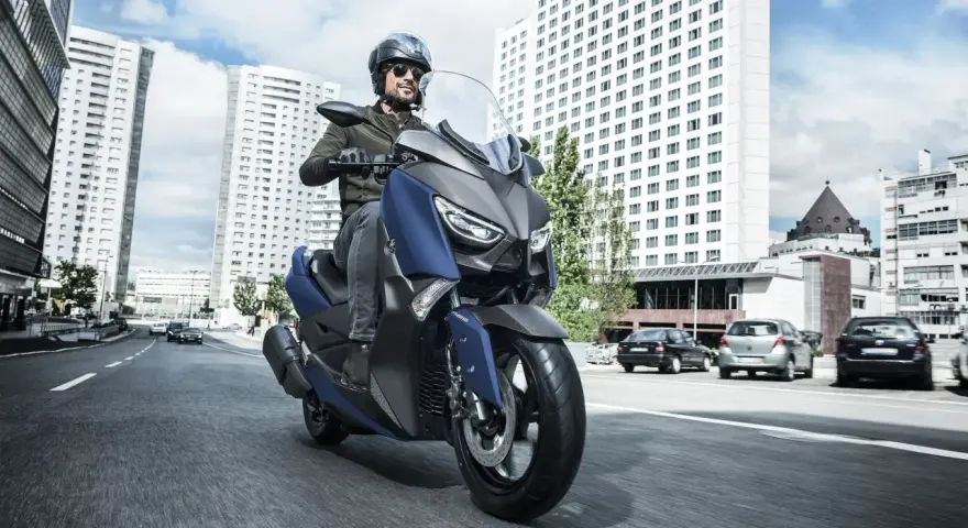 Mężczyzna w kasku i okularach przeciwsłonecznych jedzie niebieskim skuterem Yamaha X-MAX 400 po miejskiej drodze. W tle wysokie budynki i samochody.