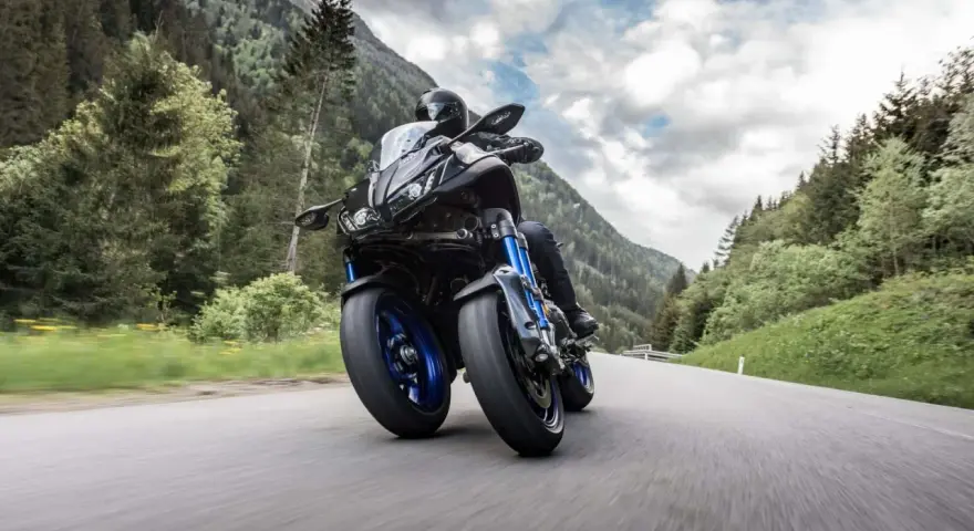 Motocyklista na Yamaha Niken pokonuje zakręt na górskiej drodze, otoczony zielenią i chmurami.