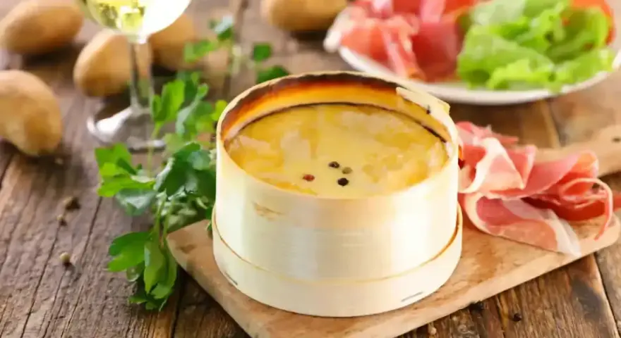 Un délicieux Mont d'Or crémeux, prêt à être dégusté avec du jambon cru, des pommes de terre et un verre de vin blanc.