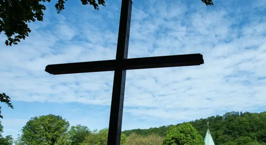 Ein großes Holzkreuz ragt vor blauem Himmel mit Wolken auf. Im Hintergrund eine Kirche und grüne Bäume. Wann ist Christi Himmelfahrt?
