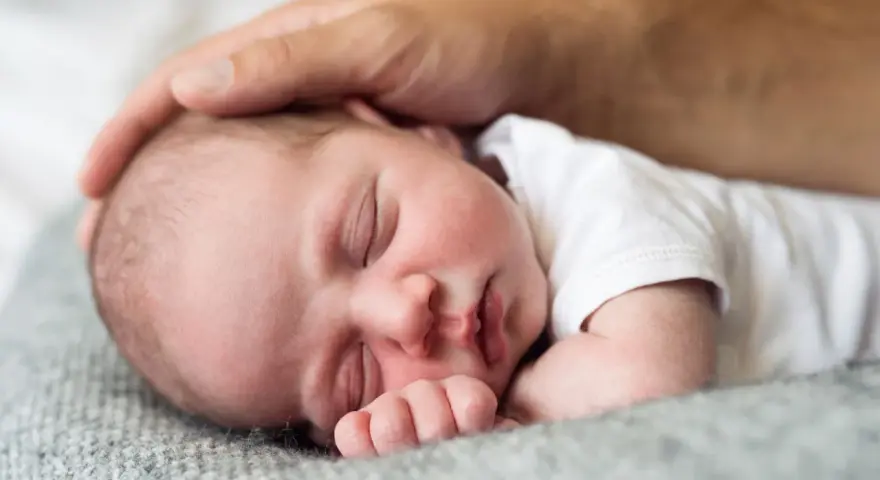 Ciepła dłoń ojca głaszcze śpiące, **2 miesięczne dziecko**. Maleństwo wtulone w miękki kocyk, wygląda na spokojne i bezpieczne.