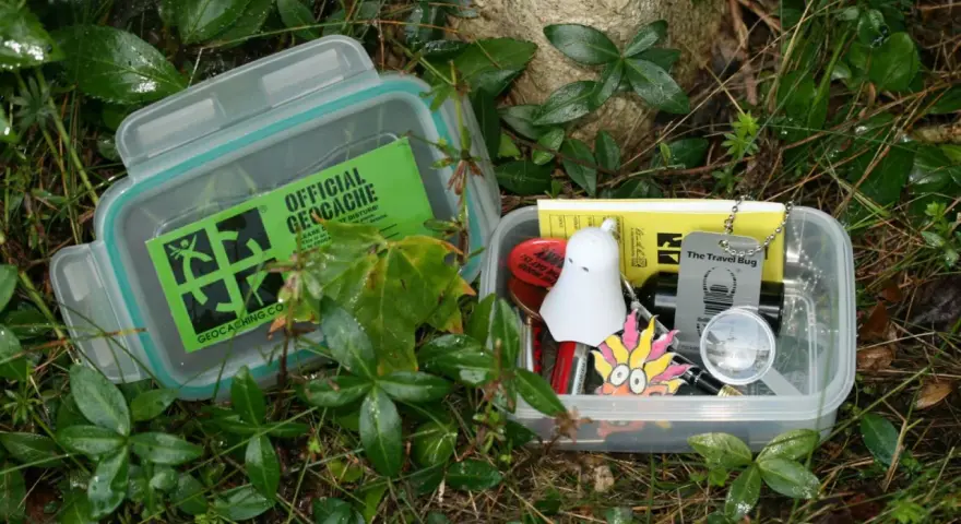 Dwa pojemniki z zawartością do geocaching co to. W jednym zielona naklejka "Official Geocache", w drugim drobne przedmioty jak duch, lupa i "Travel Bug".