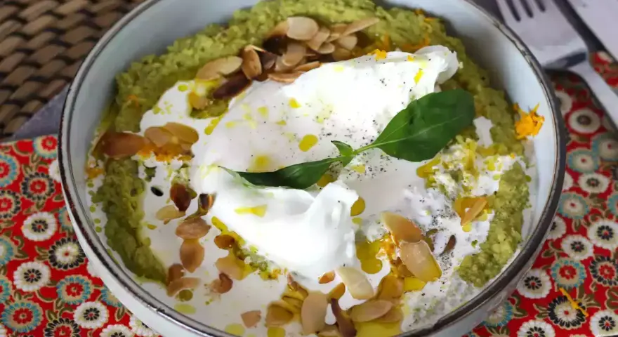 Un bol de purée verte, peut-être une sauce petit pois, garni d'un œuf poché, de crème, d'amandes effilées et d'une feuille de basilic.