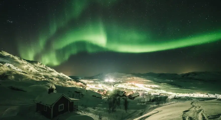 Snöklädda berg och en liten by badar i norrskenets gröna sken. En magisk natt att se norrsken.