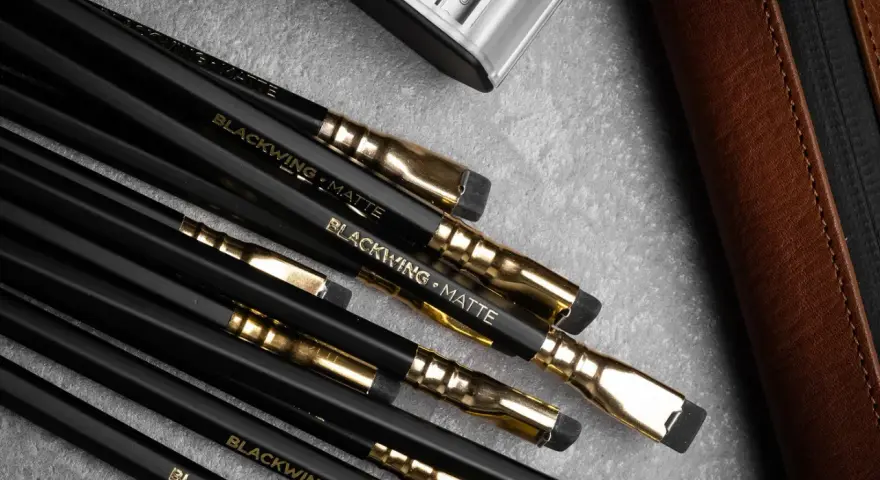 En samling svarta Blackwing-pennor med guldfattningar och suddgummin. En brun läderfodral och en metallbehållare syns också.