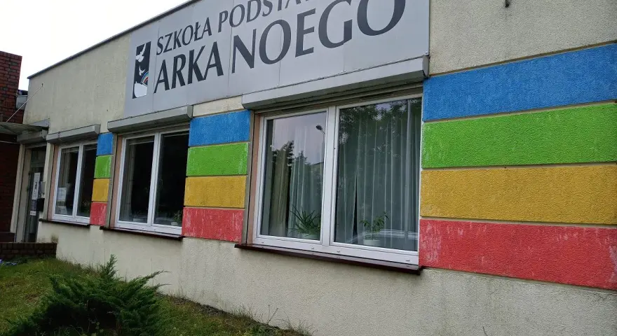 Budynek szkoły z kolorowymi pasami i napisem "SZKOŁA PODSTAWOWA ARKA NOEGO".