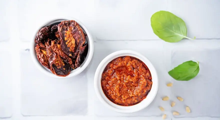 Due ciotoline bianche: una con pomodori secchi interi, l'altra con un invitante pesto di pomodori secchi. Foglie di basilico e pinoli completano la scena.