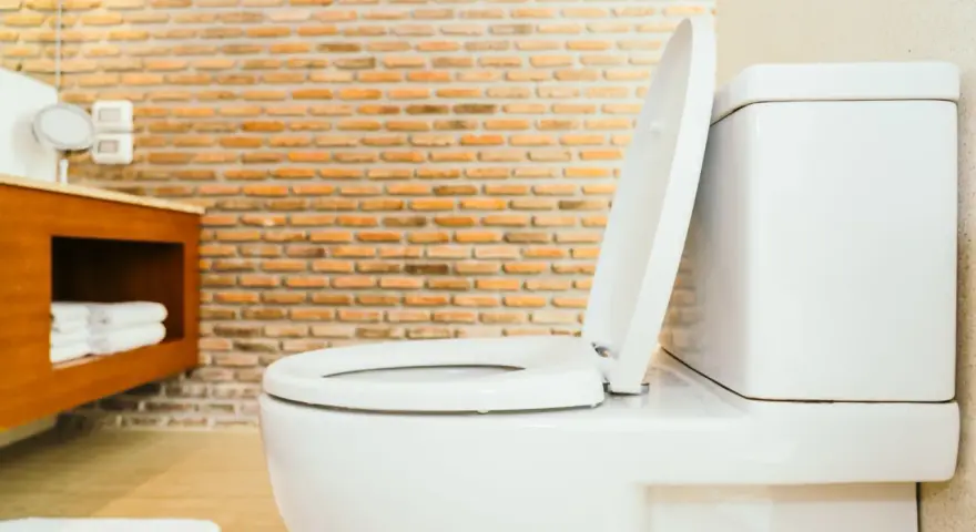 Biała miska WC z podniesioną deską, widoczna w łazience z ceglaną ścianą. Określenie jaka wysokość miski WC od podłogi jest kluczowa dla wygody.