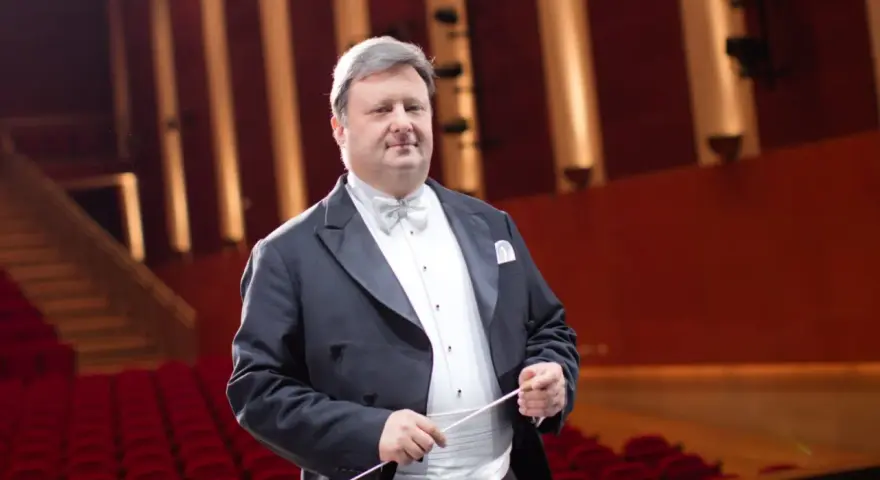 Jacek Boniecki, dyrygent w eleganckim fraku, z batutą w dłoni, na tle czerwonej widowni. Jego życiorys to historia pasji do muzyki.