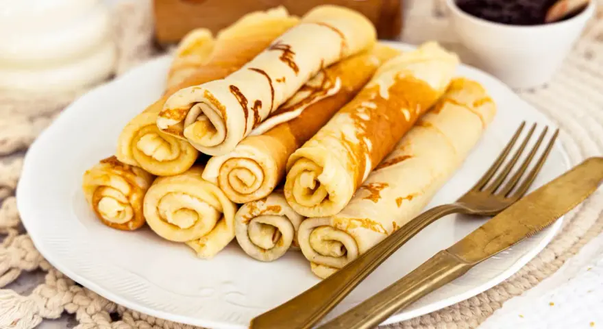Ein Teller mit goldbraunen Quark Pfannkuchen, aufgerollt und gestapelt. Daneben liegen ein goldenes Messer und eine Gabel. Im Hintergrund eine Schale Marmelade.