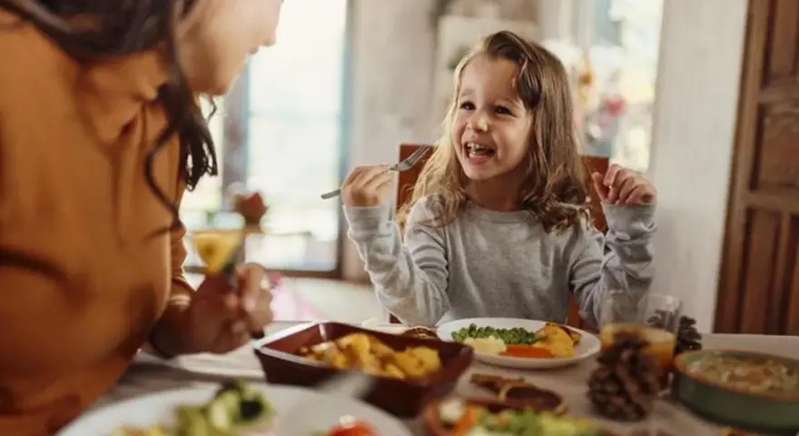 Bambina felice mangia da sola a tavola, mostrando che a che età i bambini mangiano da soli è un traguardo raggiunto con gioia.