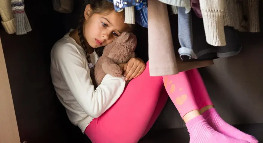Trauriges Kind versteckt sich im Schrank, umgeben von Kleidung. Die Szene erweckt den Eindruck einer Notsituation, die das Jugendamt auf den Plan rufen könnte, falls das Kind Drogenprobleme hat.