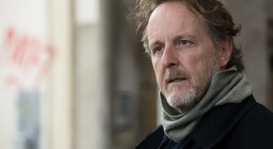 Christian Peppard, con un foulard e un cappotto scuro, guarda pensieroso verso destra.