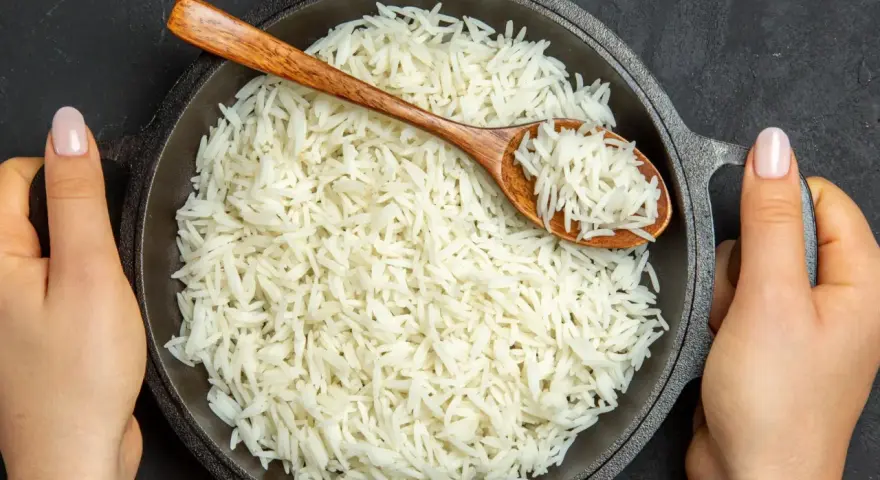 Dwie ręce trzymają okrągłą, czarną patelnię pełną puszystego ryżu basmati. Drewniana łyżka zanurzona w ryżu.