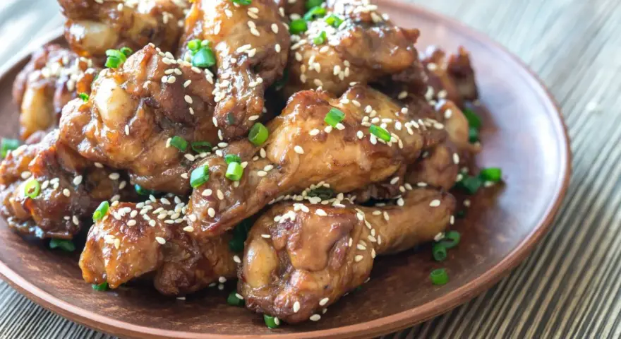 Alitas de pollo glaseadas con sésamo y cebolleta en un plato marrón.