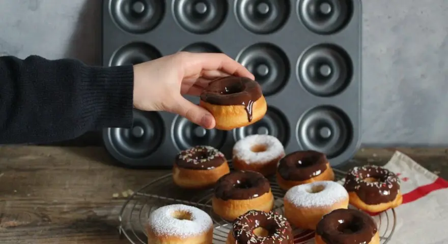 Une main tient un donut glacé au chocolat, prêt à être dégusté. Une belle collection de donuts au four, certains saupoudrés de sucre glace, d'autres de glaçage au chocolat et de vermicelles colorés, attendent sur une grille.