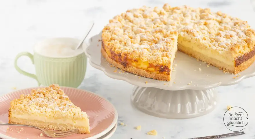 Ein saftiger Streuselkuchen mit Puddingfüllung, bestäubt mit Puderzucker, neben einer Tasse Cappuccino.