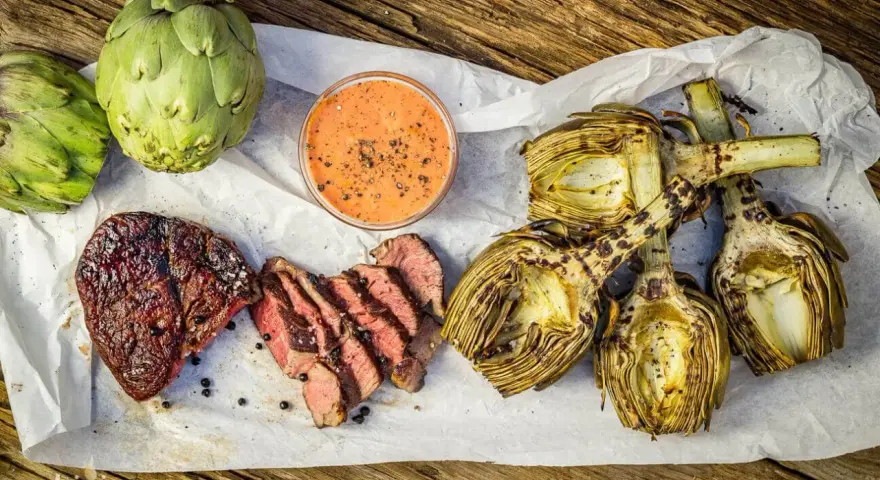 Saftiges Steak und gegrillte Artischocken mit Dip auf Holzuntergrund.