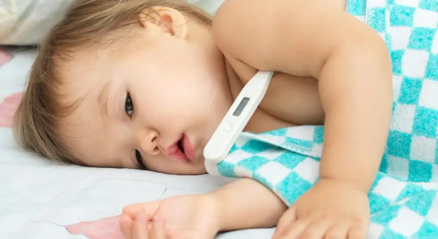 Małe dziecko z termometrem pod pachą, owinięte w kocyk. Wygląda na chore, być może to trzydniówka u dzieci.