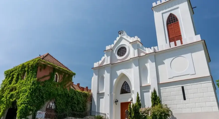Biały kościół z wieżą i zarośnięty bluszczem budynek na tle błękitnego nieba.