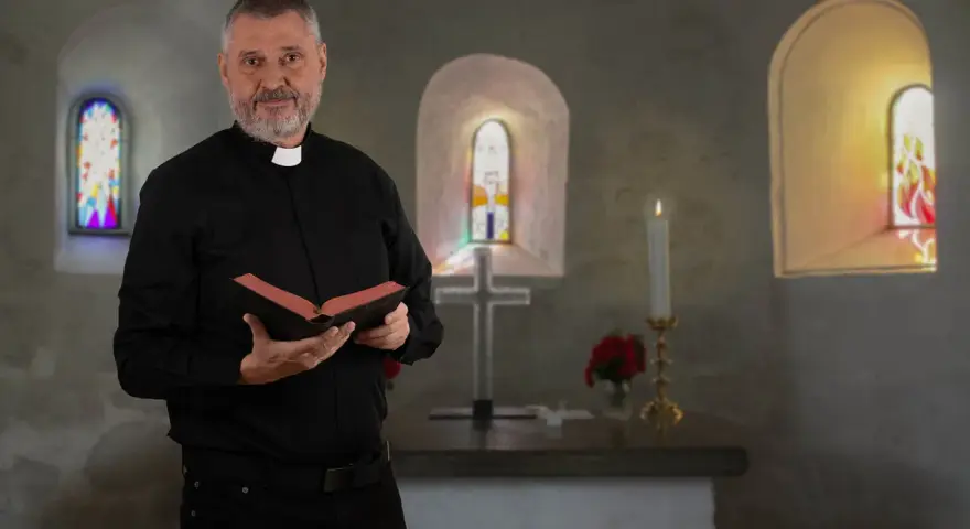 Ein Mann mit grauem Bart und Priesterkragen hält ein Buch in einer Kirche. Er hat sich entschieden, Priester zu werden.