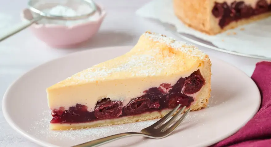 Ein saftiges Stück Kirsch Schmand Kuchen mit cremigem Belag und Kirschen auf Mürbeteigboden, bestäubt mit Puderzucker.