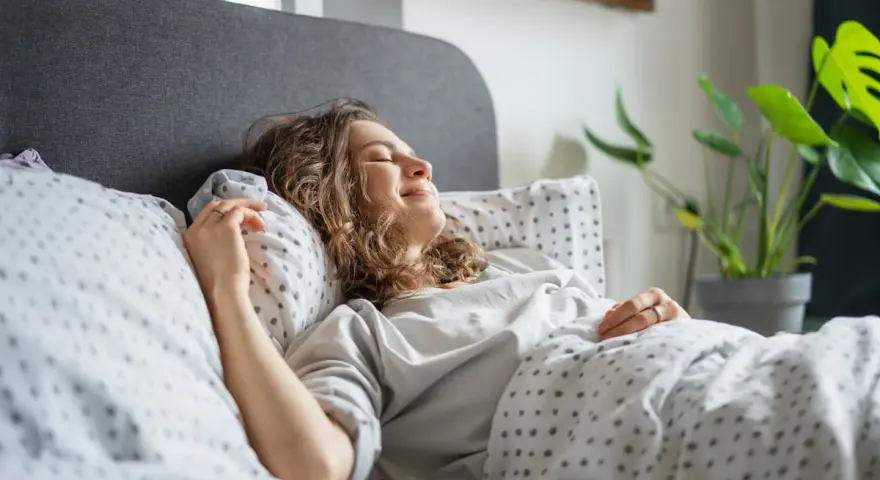 Une femme souriante se repose dans un lit douillet. Elle semble heureuse, une belle illustration de comment choisir une bonne literie pour un sommeil réparateur.