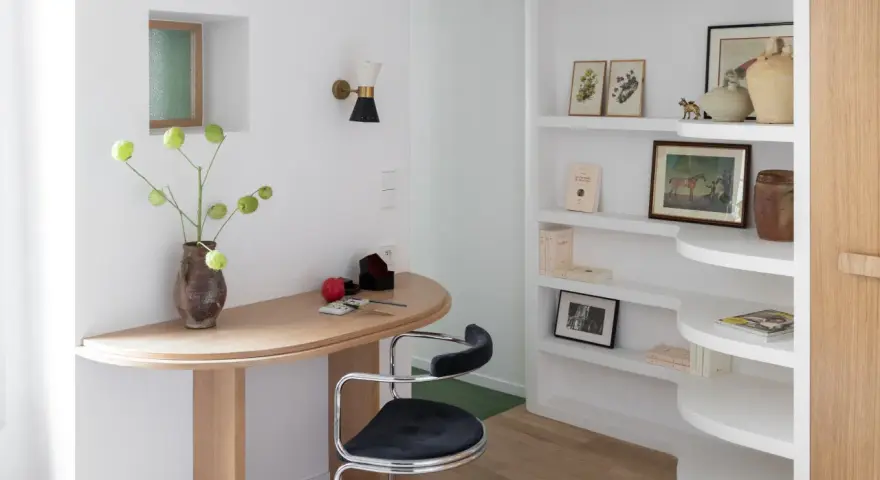 Un despacho pequeño en casa con un escritorio curvo de madera, una silla de terciopelo azul y estanterías blancas llenas de libros y objetos decorativos.