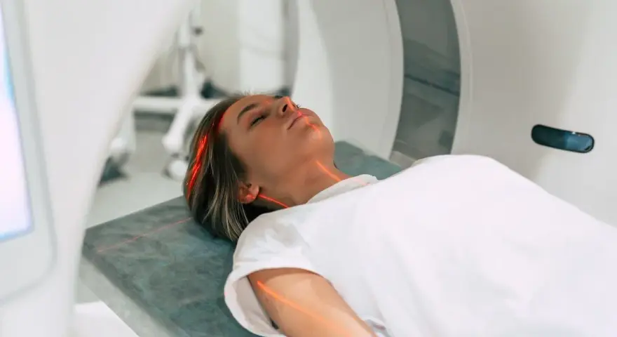 Kobieta leży na łóżku w aparacie do rezonansu magnetycznego. Czerwone linie lasera wskazują obszar badania. To właśnie mri co to za badanie?