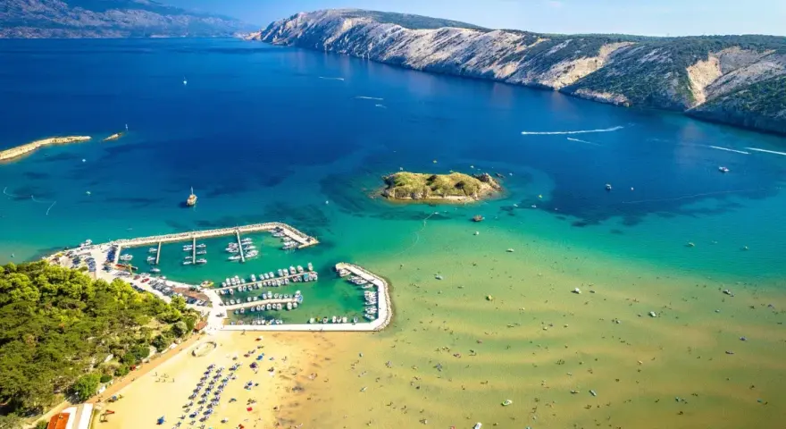 Piaszczyste plaże w Chorwacji, z turkusową wodą, łodziami w marinie i ludźmi cieszącymi się słońcem.