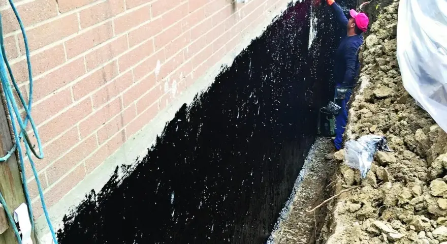 Prace przy izolacji fundamentów w starym domu. Pracownik nakłada czarną masę izolacyjną na ścianę fundamentową.