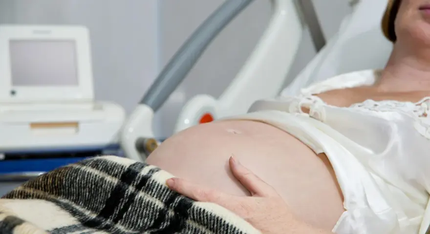 Brzuch kobiety w ciąży, ręka na brzuchu, monitor KTG i łóżko szpitalne.