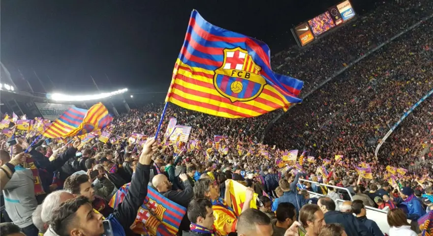 Tłum kibiców na stadionie unosi flagi z herbem FC Barcelona. Flaga Katalonii powiewa dumnie nad głowami fanów.