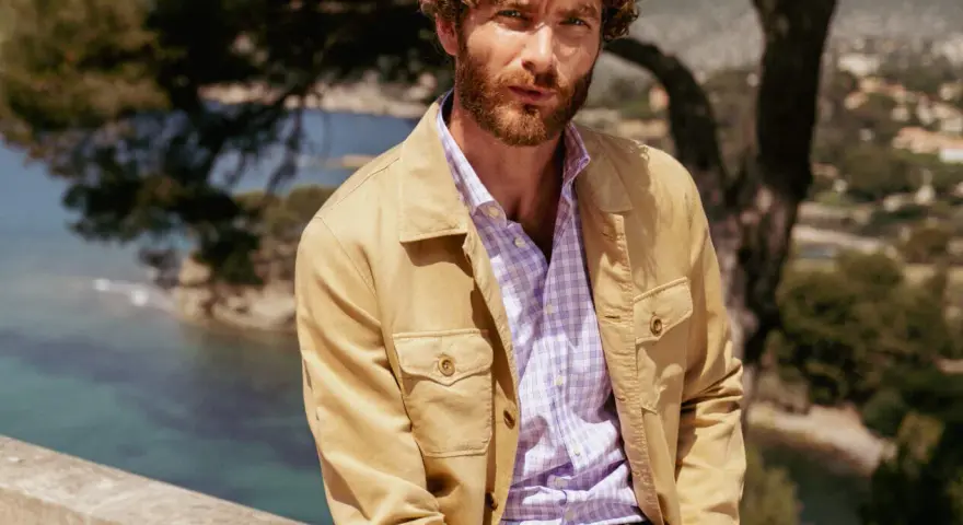 Un uomo con barba e capelli ricci indossa una camicia a quadri viola e una giacca color sabbia. Ottima idea su cosa mettere sopra la camicia uomo per un look casual chic.