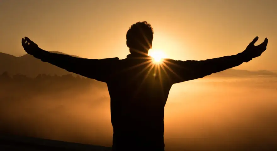 Silhouette eines Mannes mit ausgebreiteten Armen im Sonnenaufgang. Die Sonne bricht durch seine Silhouette und wirft Strahlen. Ein Symbol für Hoffnung und wie wird man Christ.