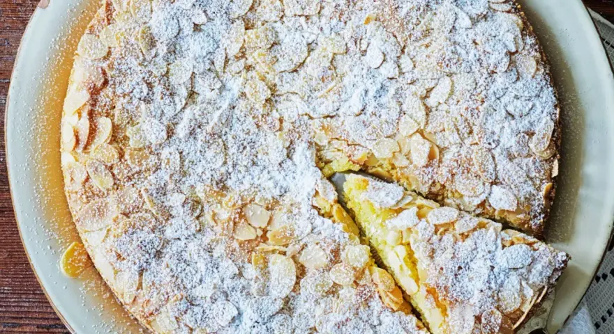 En saftig mandelkaka med citron, toppad med flagad mandel och pudrad florsocker. En bit är uppskuren.