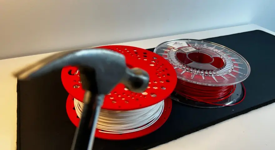 Młotek zbliża się do szpul z filamentem, testując wytrzymałość materiałów. Biały i czerwony filament czekają na próbę.