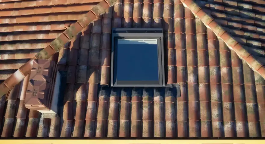 Un tetto in tegole rosse, con una finestra da tetto moderna e un comignolo. Le tegole mostrano segni del tempo, con muschio e licheni che aggiungono carattere.