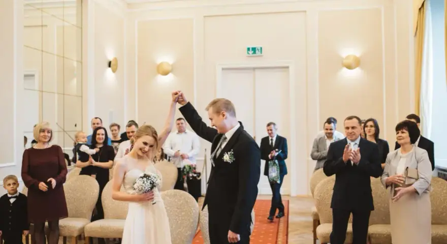 Para młoda świętuje swój ślub cywilny, trzymając się za ręce. W tle widać gości i rodzinę.