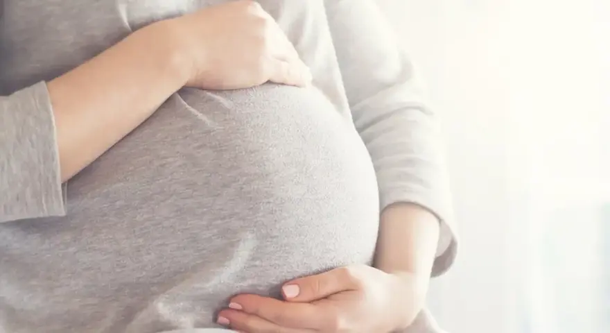 Brzuszek w 6 miesiącu ciąży, otulony szarym materiałem. Dłonie delikatnie go obejmują, tworząc ciepły krąg.