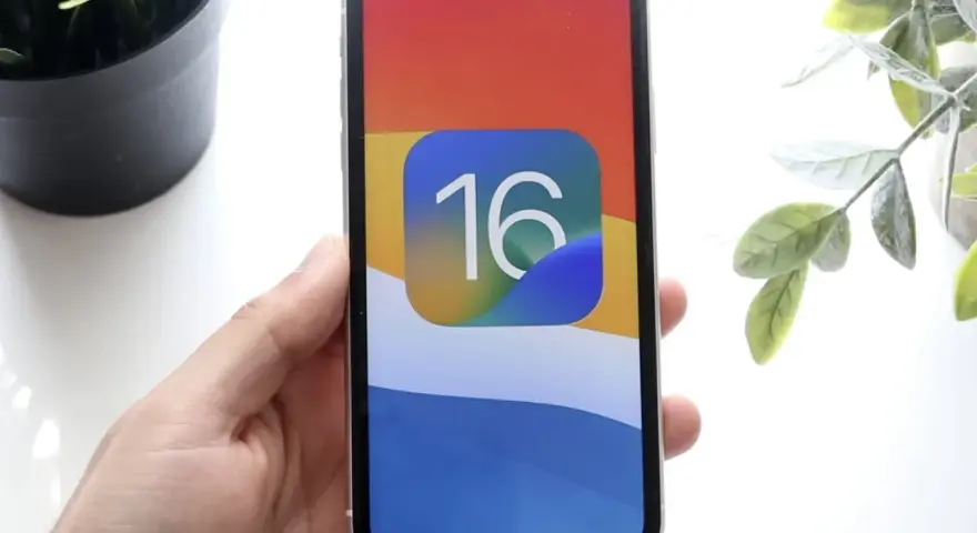 Dłoń trzyma iPhone'a z ikoną iOS 16 na kolorowej tapetcie.